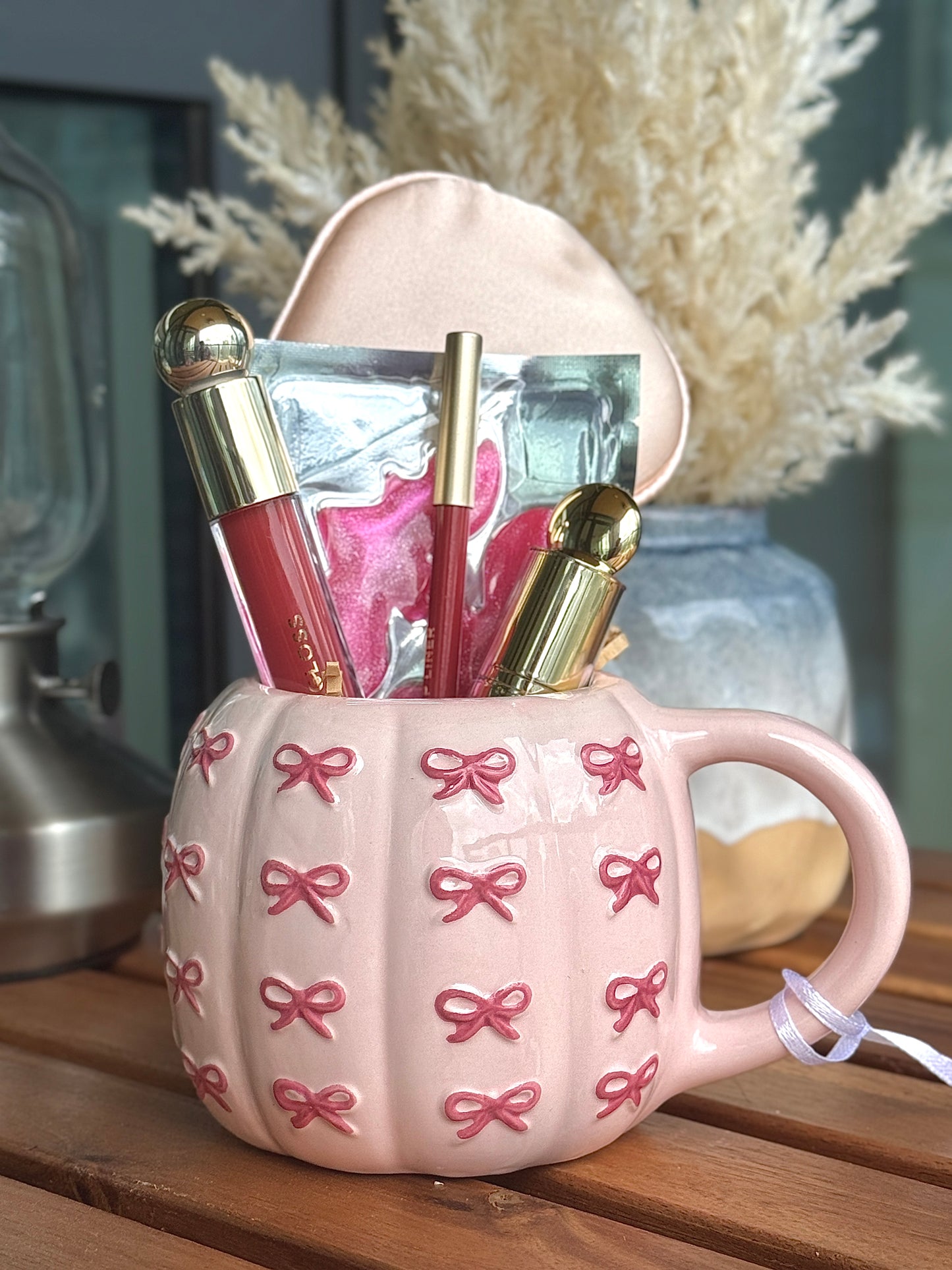 Pink mug with bow pattern filled with lipstick, lip liner, gloss, satin eye mask and under eye patch.